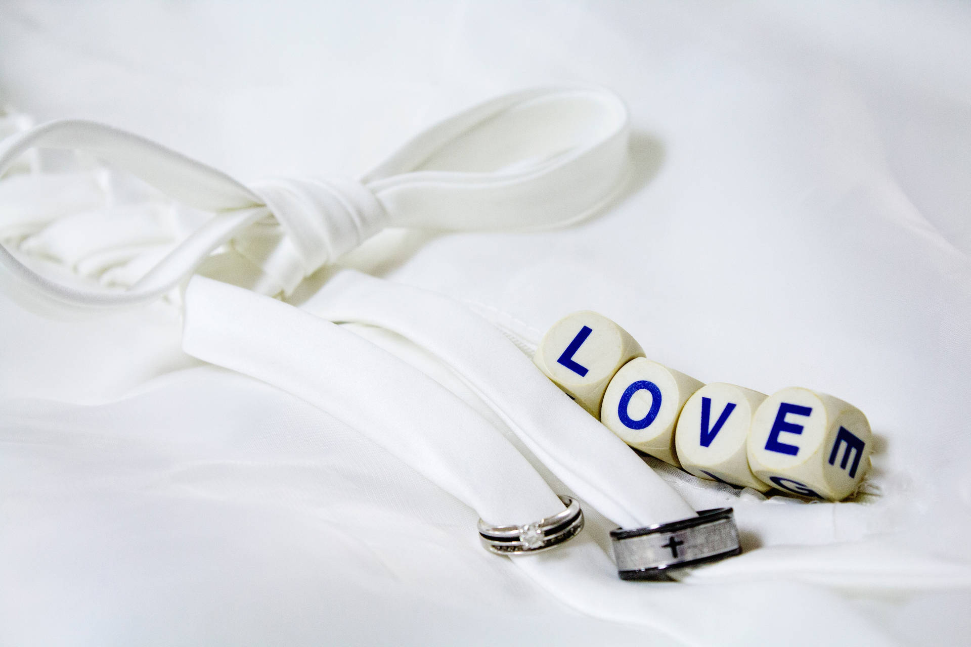 ข้อความ "Love" เขียนบนลูกเต๋าสีขาวสี่ลูก ข้างๆ แหวนแต่งงานสีเงินและสีดำสองวงผูกด้วยริบบิ้นสีขาว วางบนผ้าสีขาว — ความรัก วอลเปเปอร์