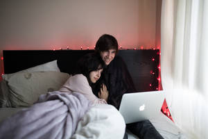 คู่รักบนเตียงใช้ MacBook Air ข้างหน้าต่างที่มีม่านสีขาว — MacBook Pro วอลเปเปอร์