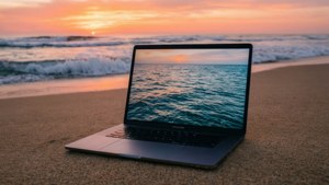 MacBook ที่สงบนิ่งบนชายหาดในช่วงพระอาทิตย์ขึ้นพร้อมโทนสีของมหาสมุทร
